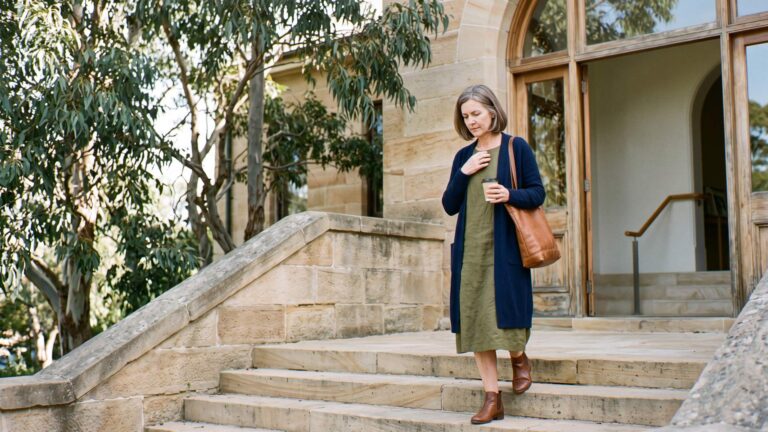 A woman in her early fifties pauses on sandstone steps under gum trees, one hand to her chest, gathering herself before the day